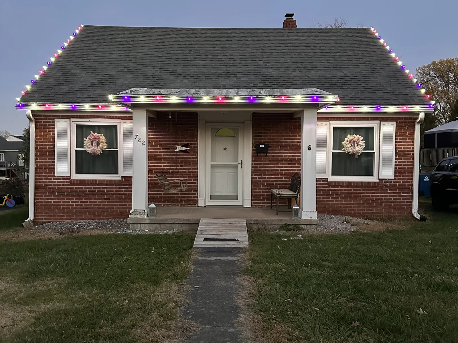 A small brick home in multicolor lights, with a glowing front porch and a wreath in each front window